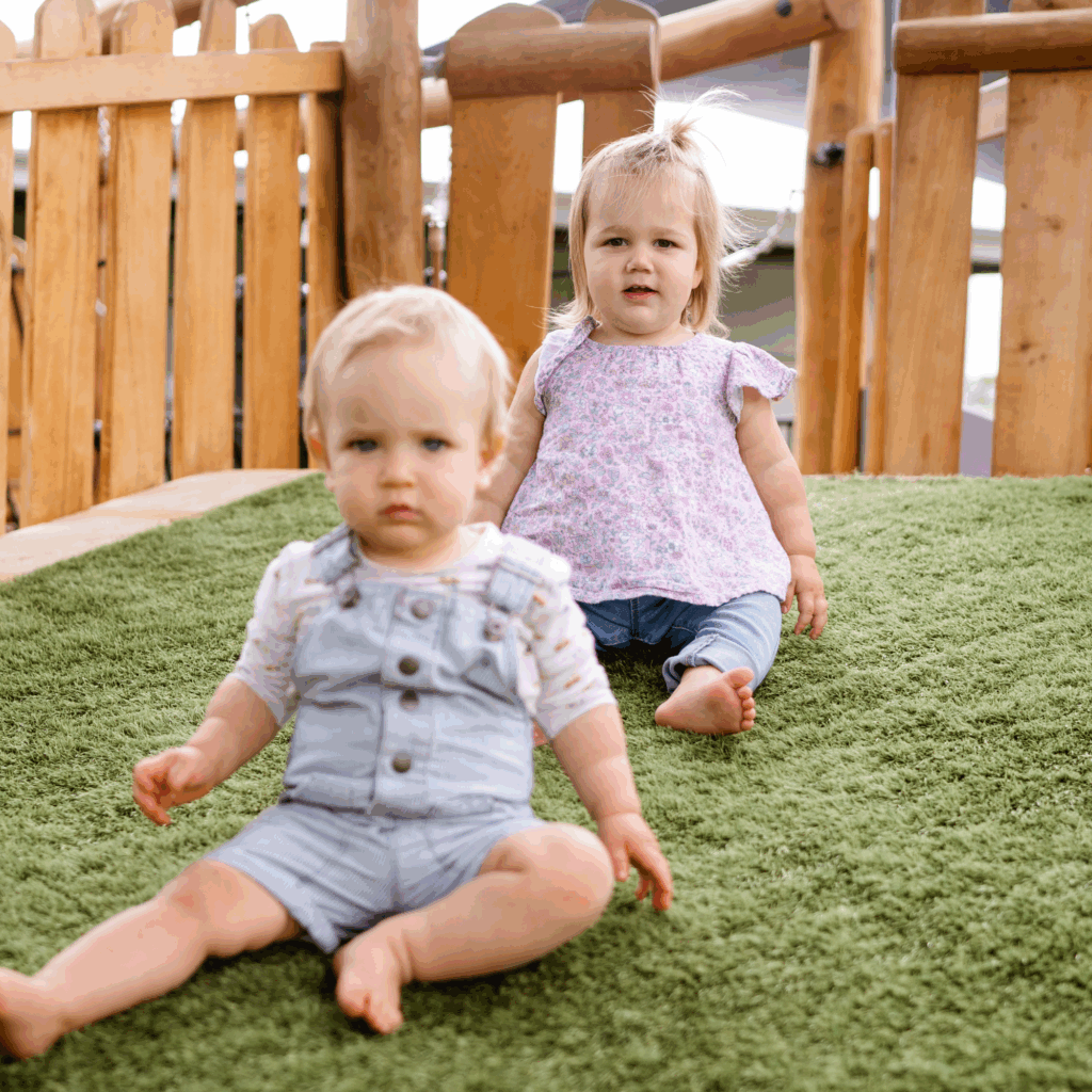 A baby boy and baby girl sat on a hill in the outdoor play space of a childcare centre